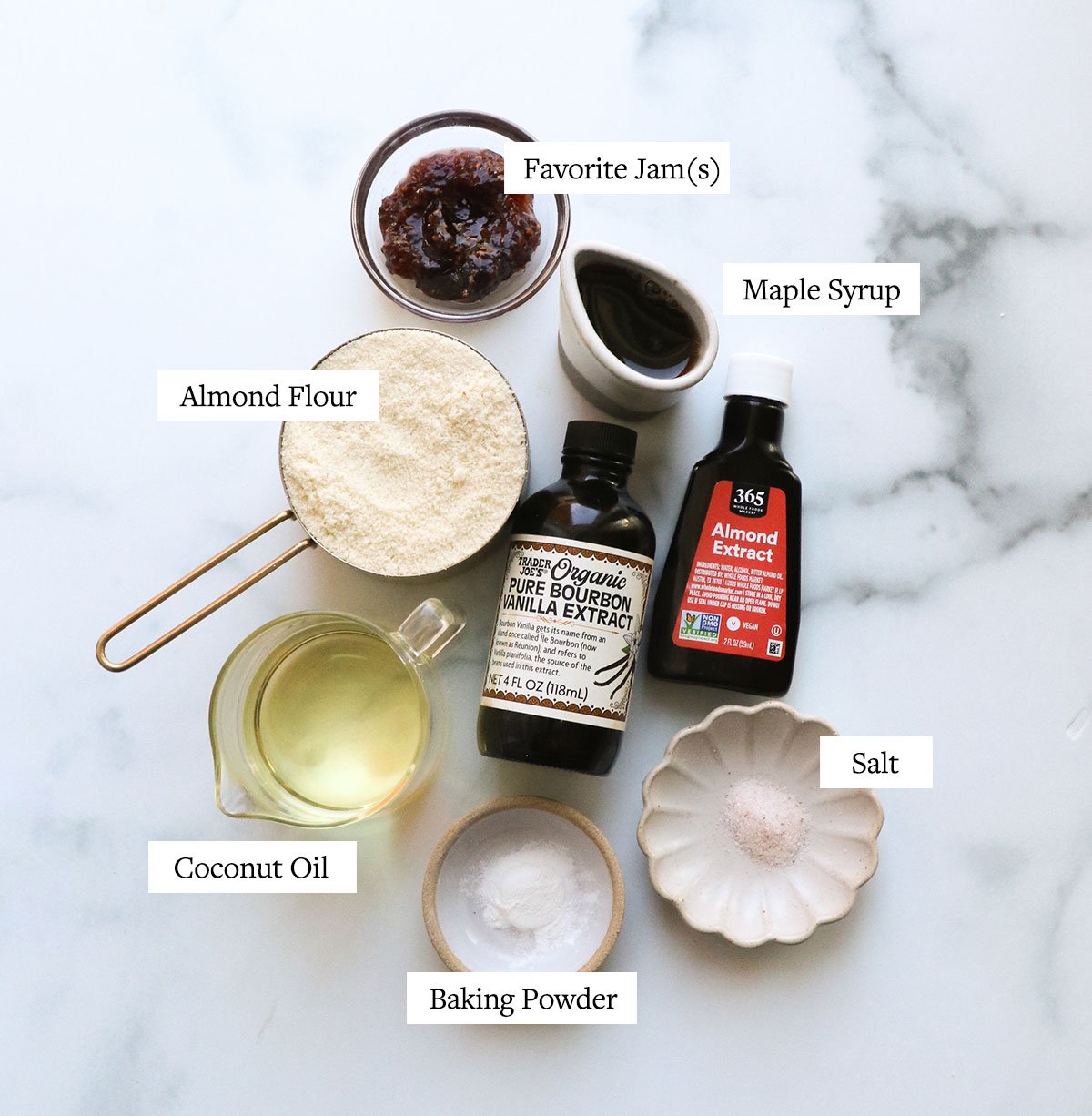 almond flour, coconut oil, baking powder, jam, and vanilla extract labeled on a marble surface.