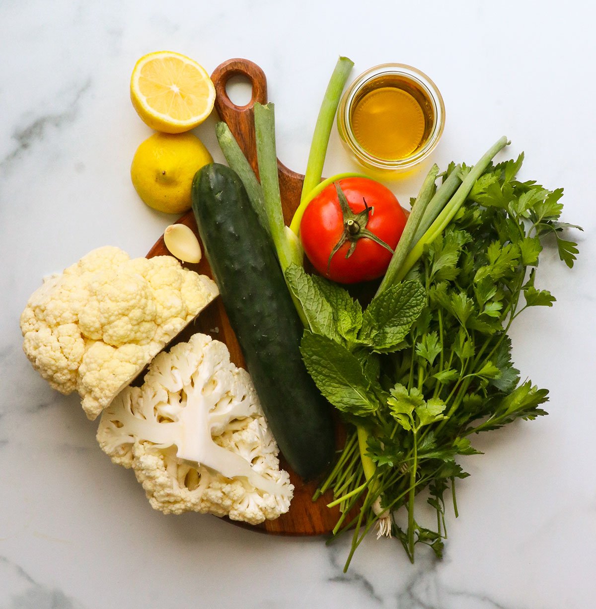 cauliflower, green onion, tomato, lemon, cucumber, and herbs on a board.