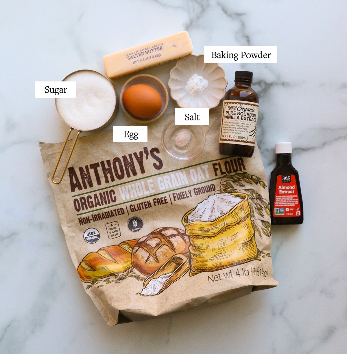 bag of oat flour near a cup of sugar, egg, butter, baking powder, vanilla and almond extract bottles.