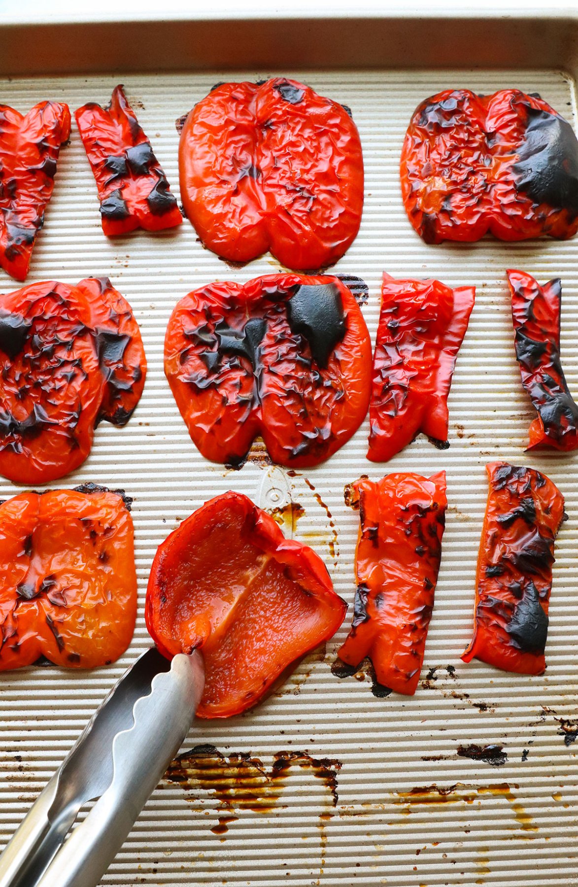 roasted red peppers on a baking sheet with one getting flipped over by a pair of tongs. 