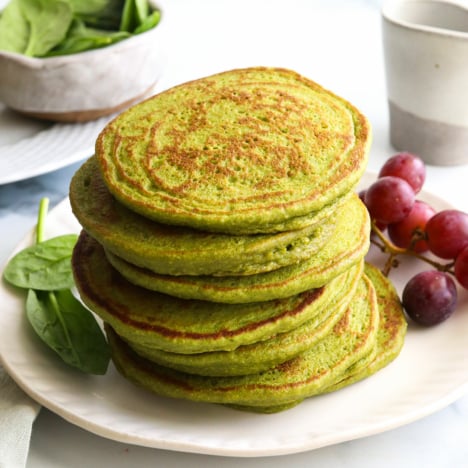 spinach pancakes stacked on a white plate by red grapes.