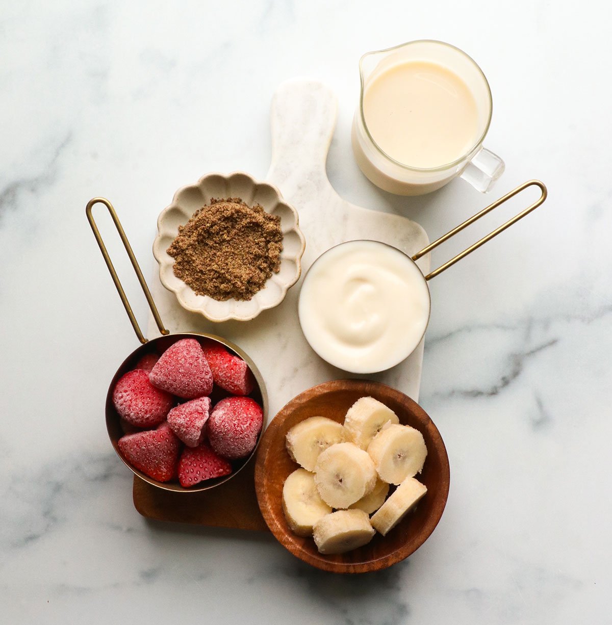 yogurt, frozen strawberries, sliced frozen bananas, milk, and flax seeds on a white surface.