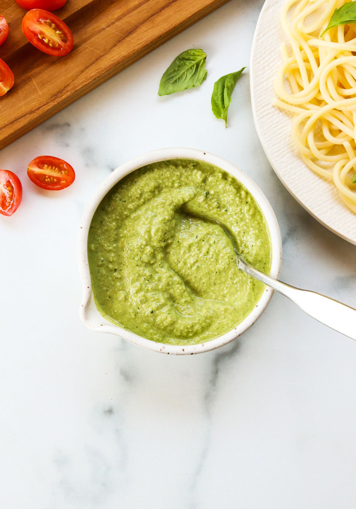 zucchini pesto in a white stone bowl with spoon next to a plate of pasta.